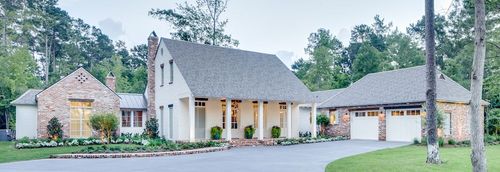 A light-colored house with a brick section and a two-car garage, surrounded by green trees on a sunny day.