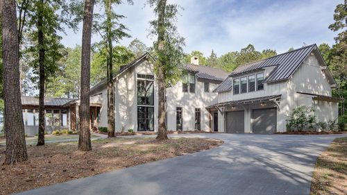 A two-story white house with a dark metal roof, set in a wooded area with a paved driveway.