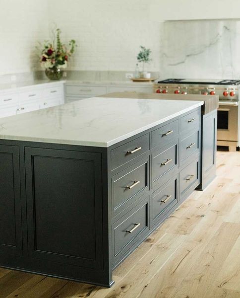A large, dark kitchen island with a white marble countertop stands in a bright kitchen with light wooden floors.