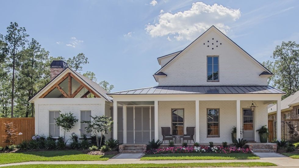A white two-story farmhouse-style home with a screened porch, metal roof, and front garden under a clear blue sky.
