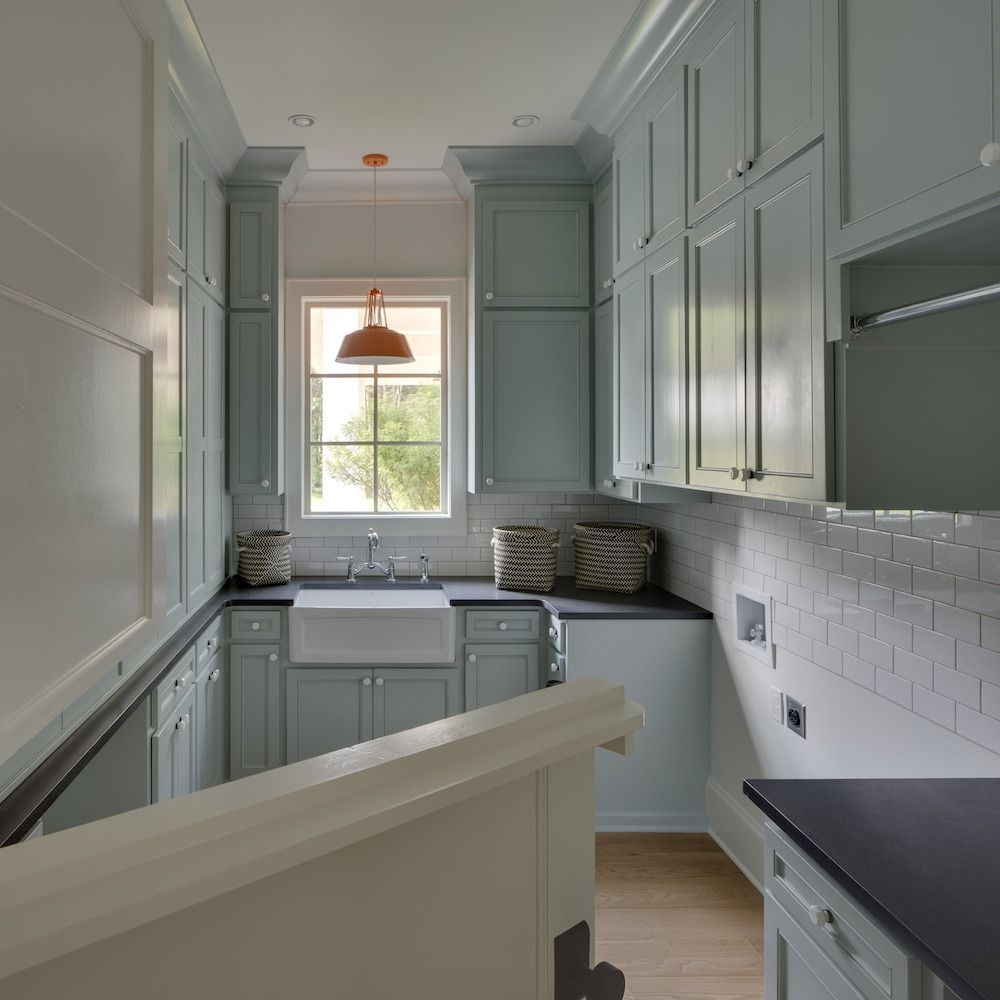 A blue-gray kitchen featuring a farmhouse sink under a window, white subway tile backsplashes, and dark countertops.