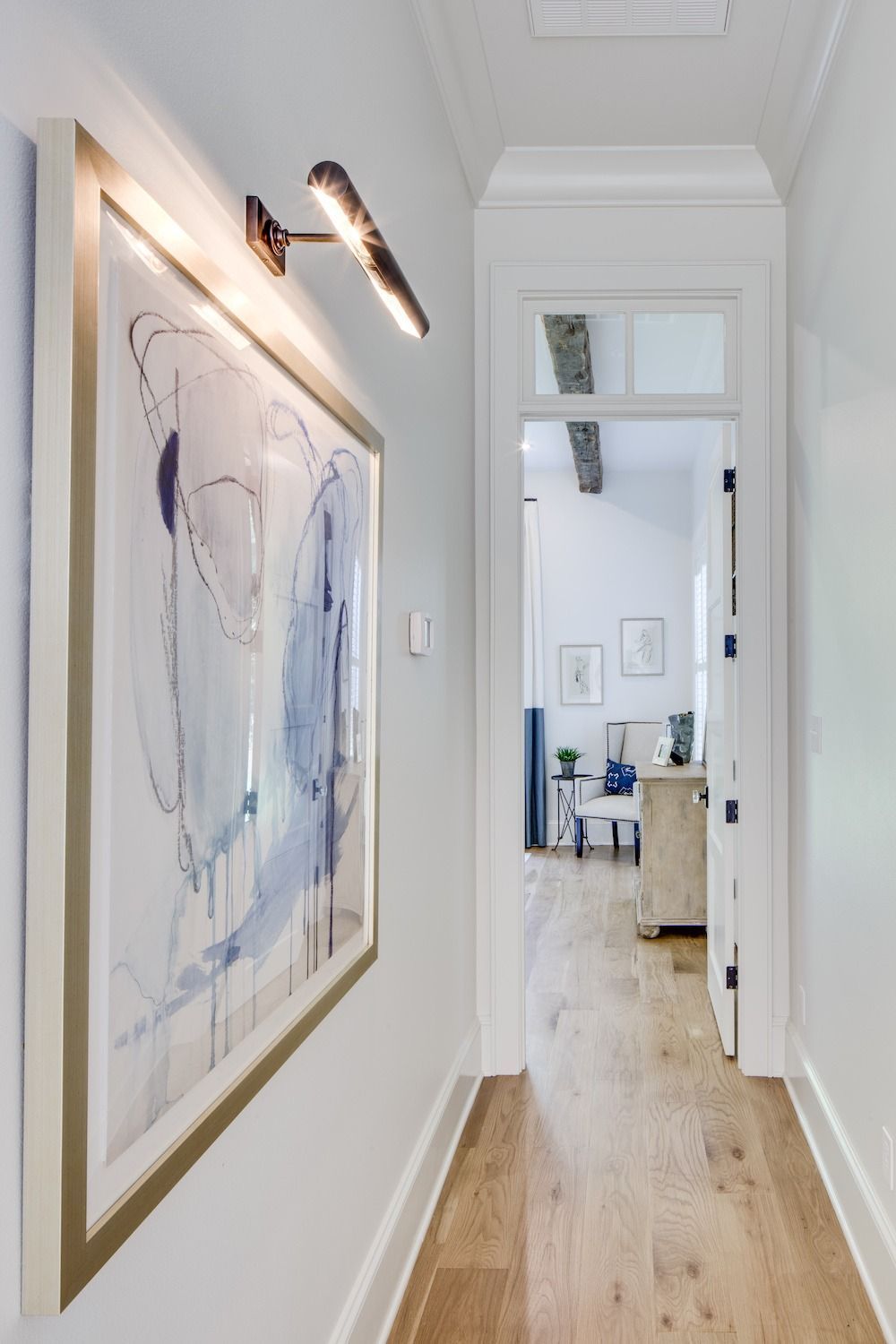 A hallway with light wood floors, a framed blue abstract art piece on the left, and a lighted entry to an adjacent room.