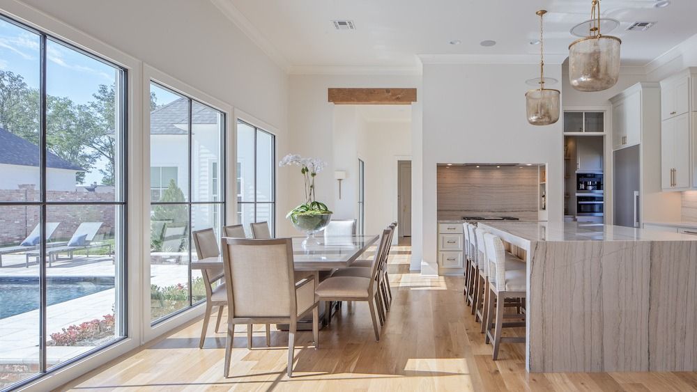 A modern, sunlit dining and kitchen area with a wooden table, beige chairs, a large kitchen island, and view of a pool.