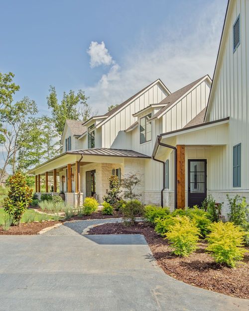 A modern white farmhouse with board-and-batten siding, stone accents, and a covered porch, set against a blue sky.
