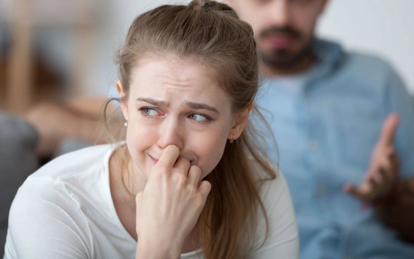 A woman is crying while sitting on a couch with a man behind her.
