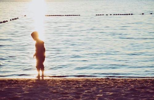 A young boy is standing on the beach looking at the water.