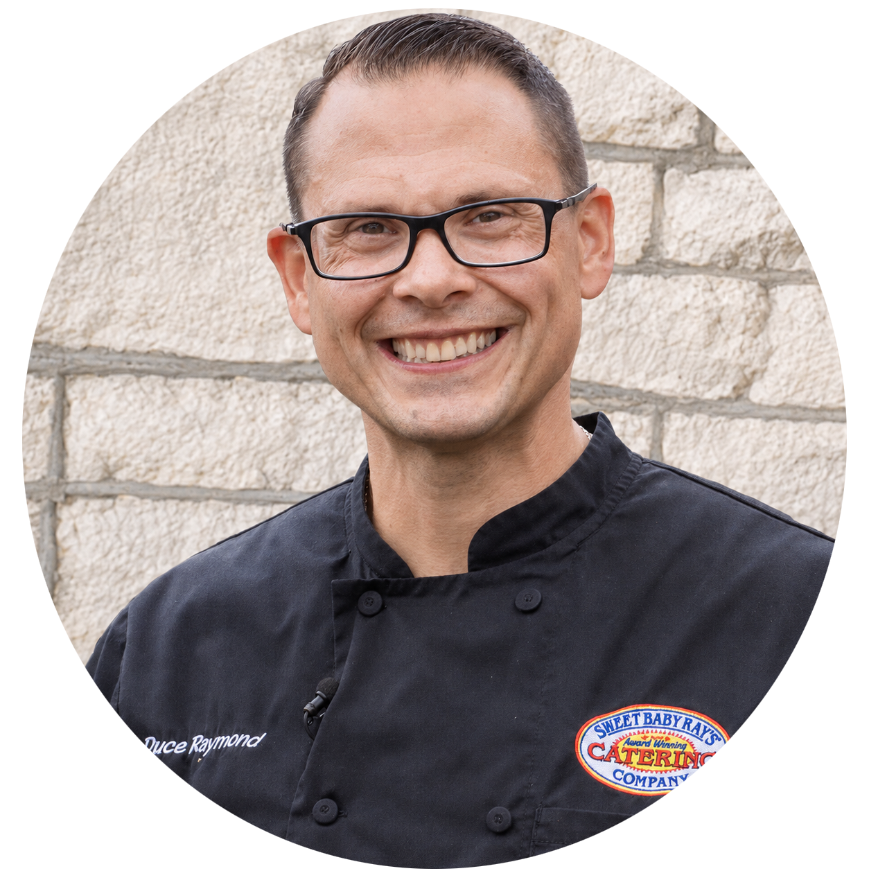 A man in a chef 's uniform is smiling in front of a brick wall