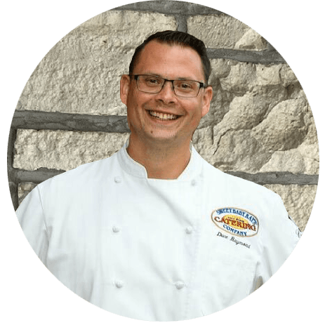 A man in a chef 's uniform is smiling in front of a stone wall.