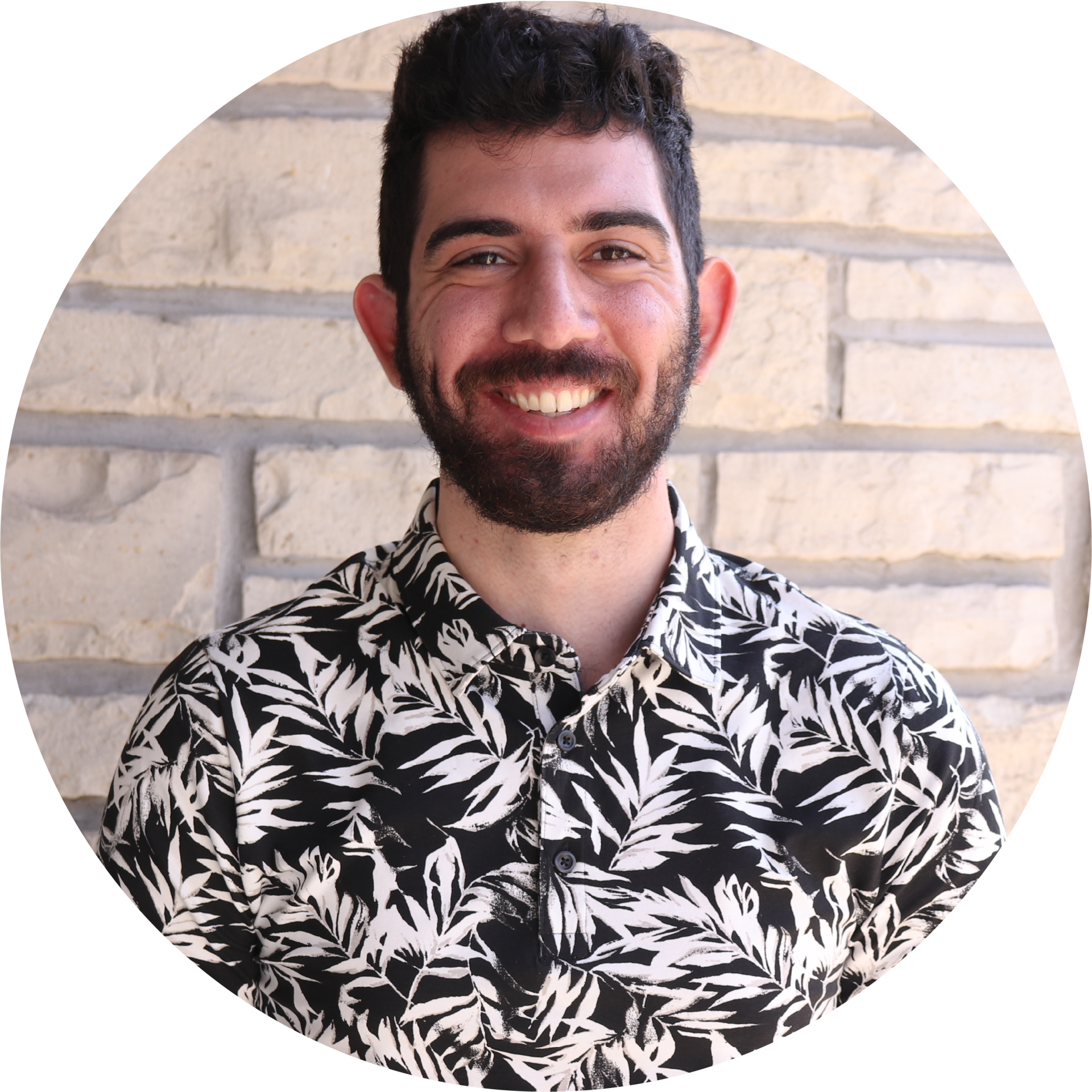 A man with a beard wearing a black and white shirt is smiling in front of a brick wall.