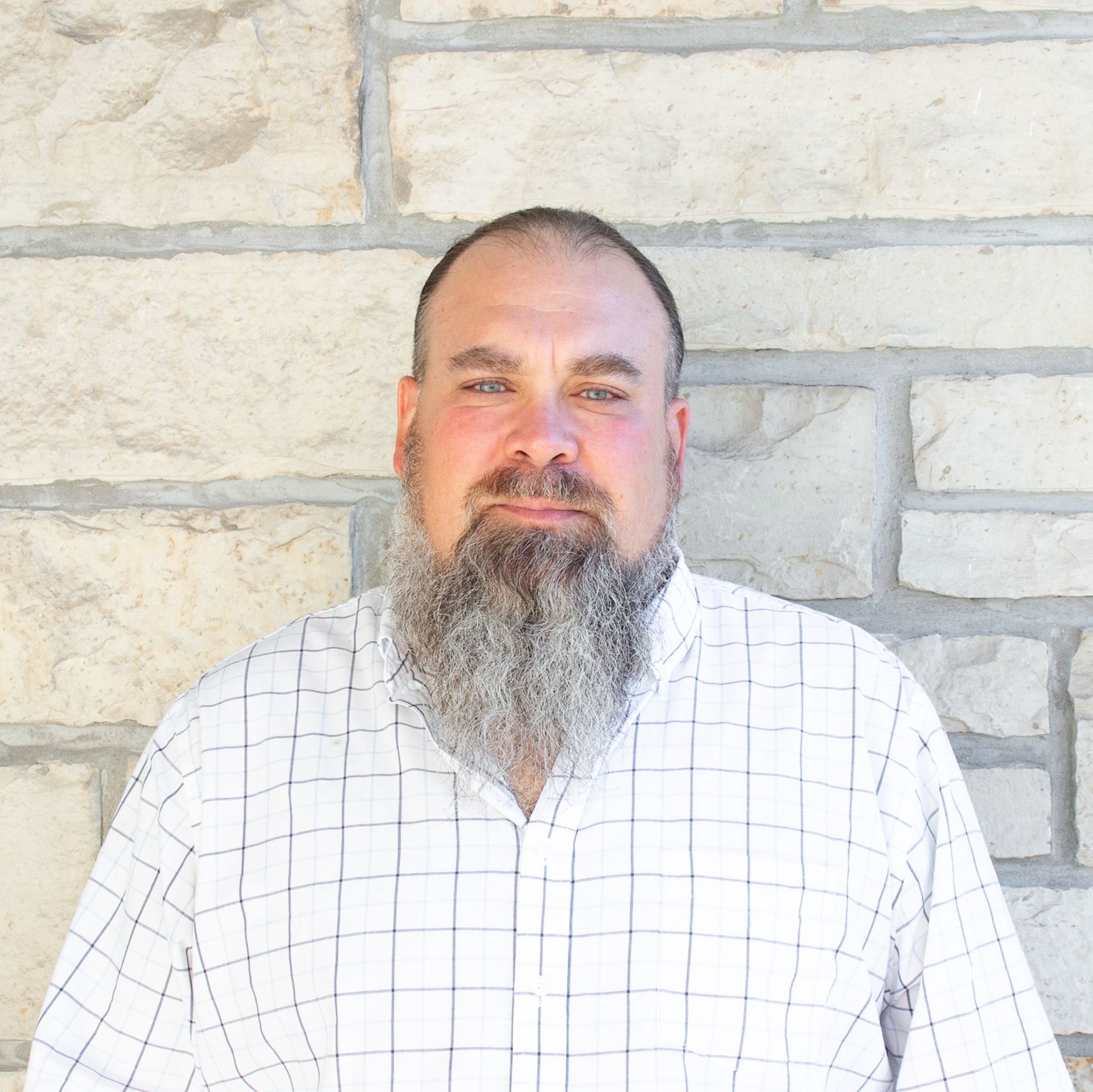 A man with a beard wearing a plaid shirt is standing in front of a brick wall.