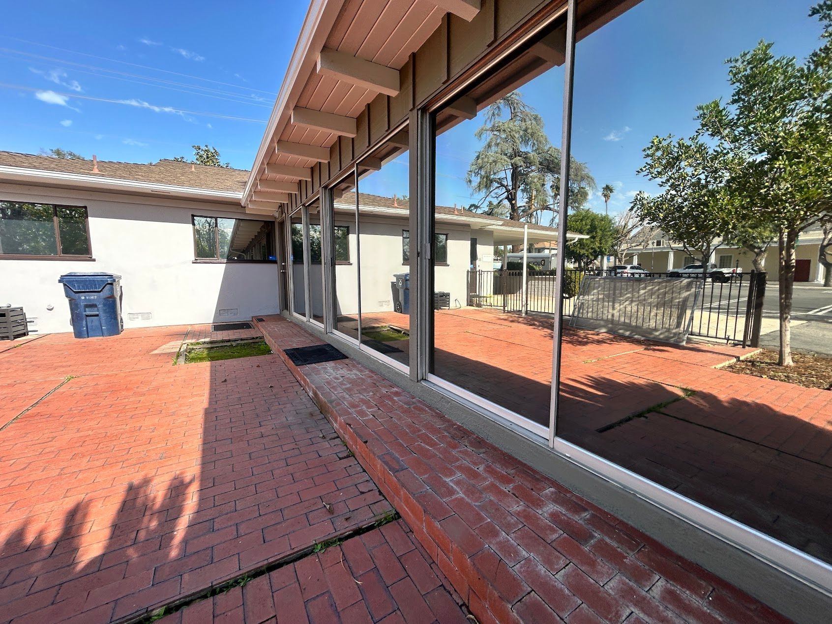 Red brick patio beside a glass-walled building, with a covered walkway and trees beyond.