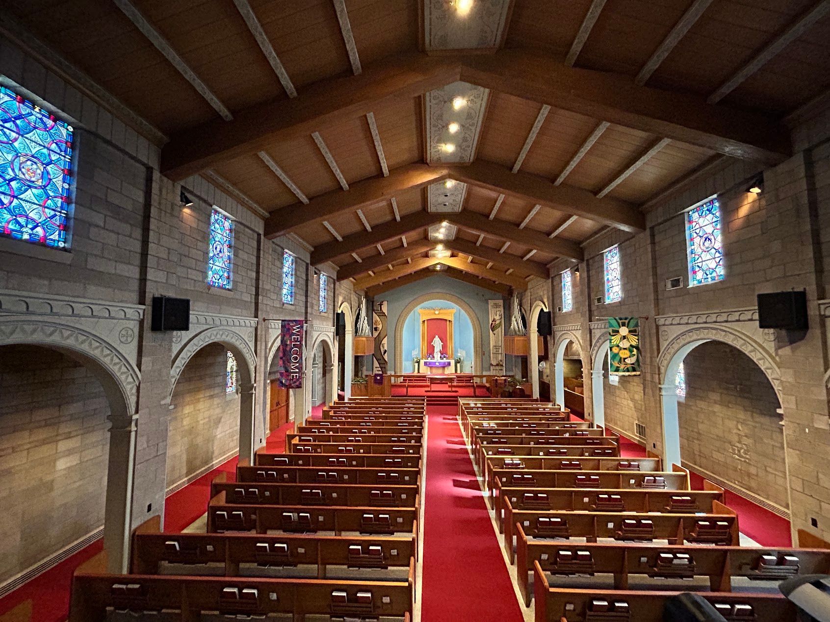 Empty church interior with red carpet aisle, wooden pews, stained glass windows, and altar at the far end