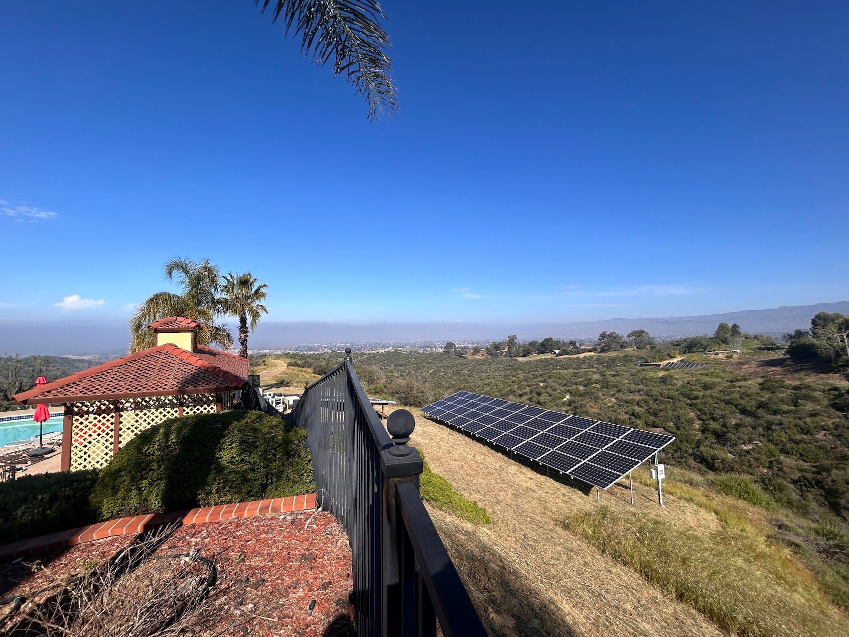 Solar panels on a hillside beside a black fence, overlooking a coastal landscape under a clear blue sky.