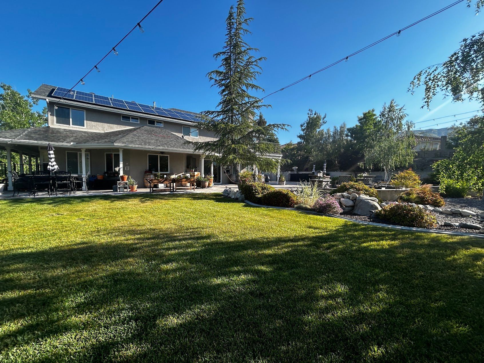 Sunny backyard with a patio, lush green lawn, trees, and flower beds beside a house