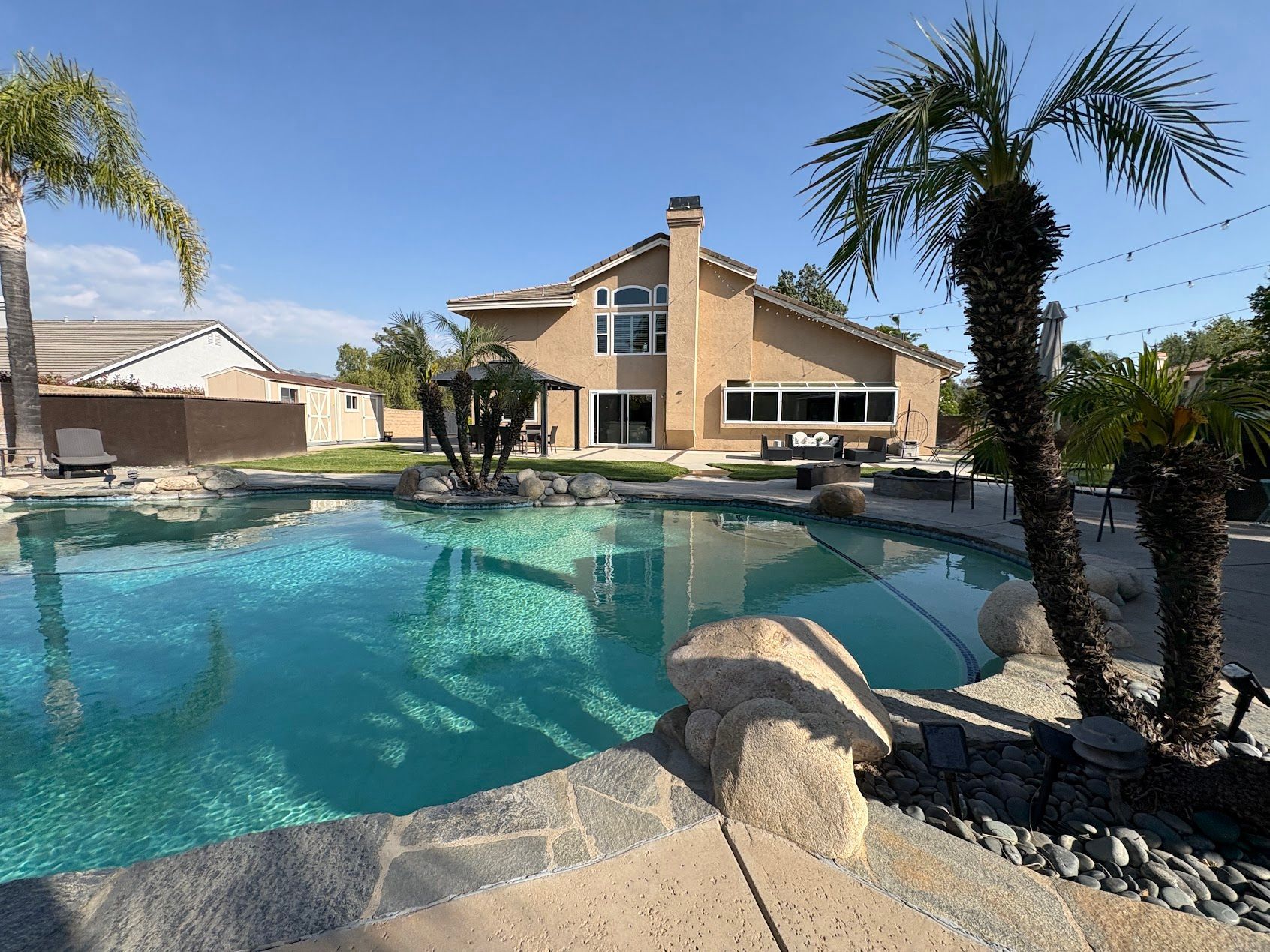 Backyard pool with a two-story house, palm trees, and bright blue water under a clear sky