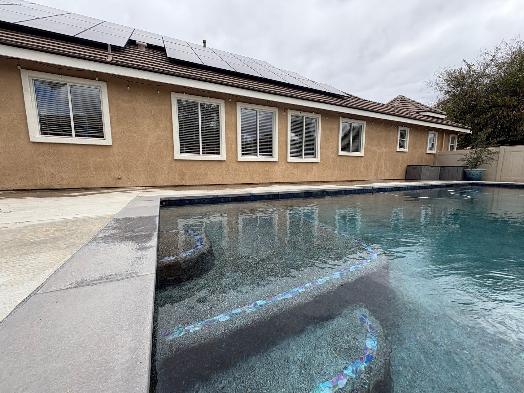 Backyard swimming pool beside a tan house with white shutters and a tiled roof