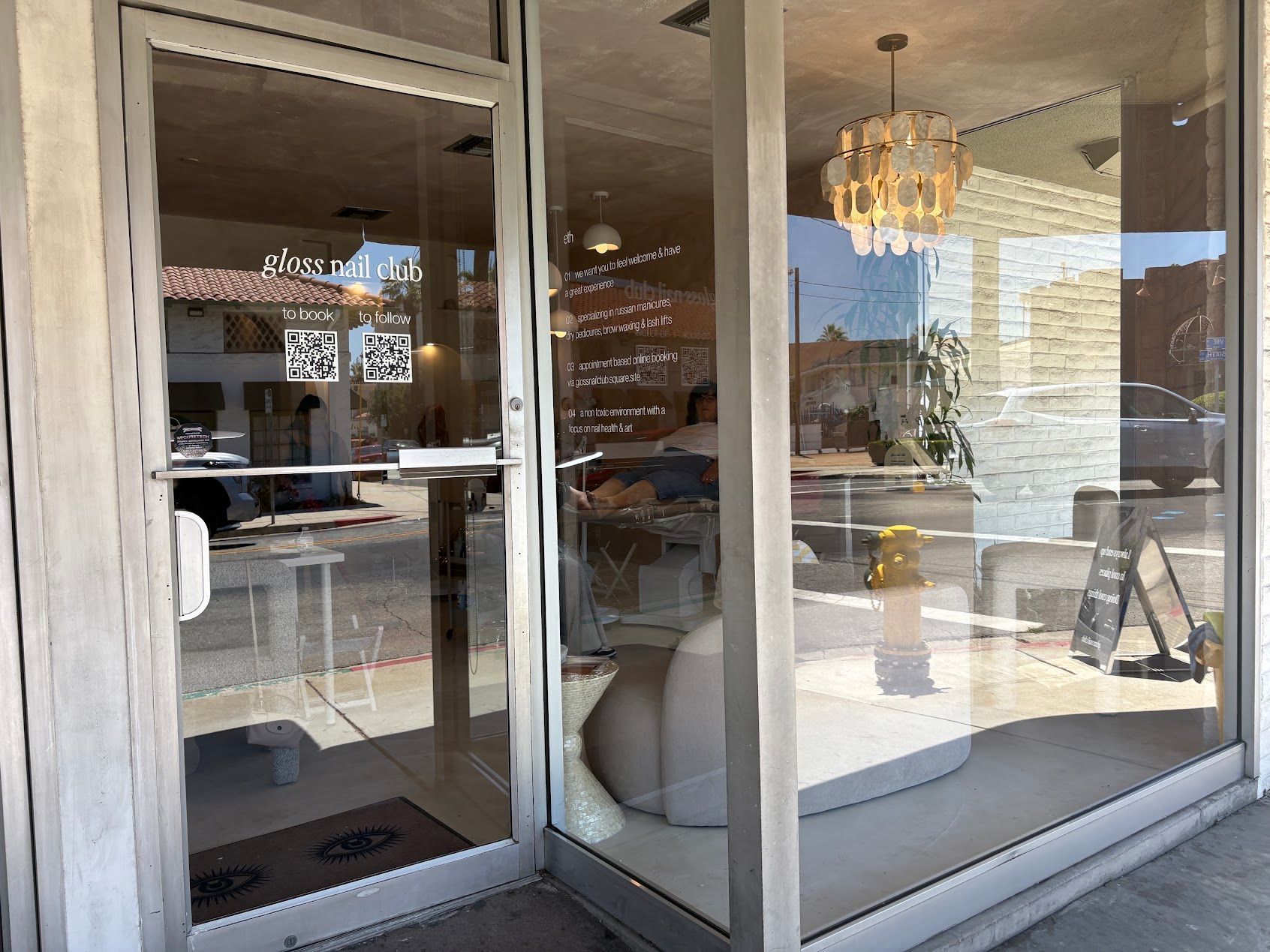 Glass storefront with interior seating visible, including a white chair and chandelier in a bright cafe.