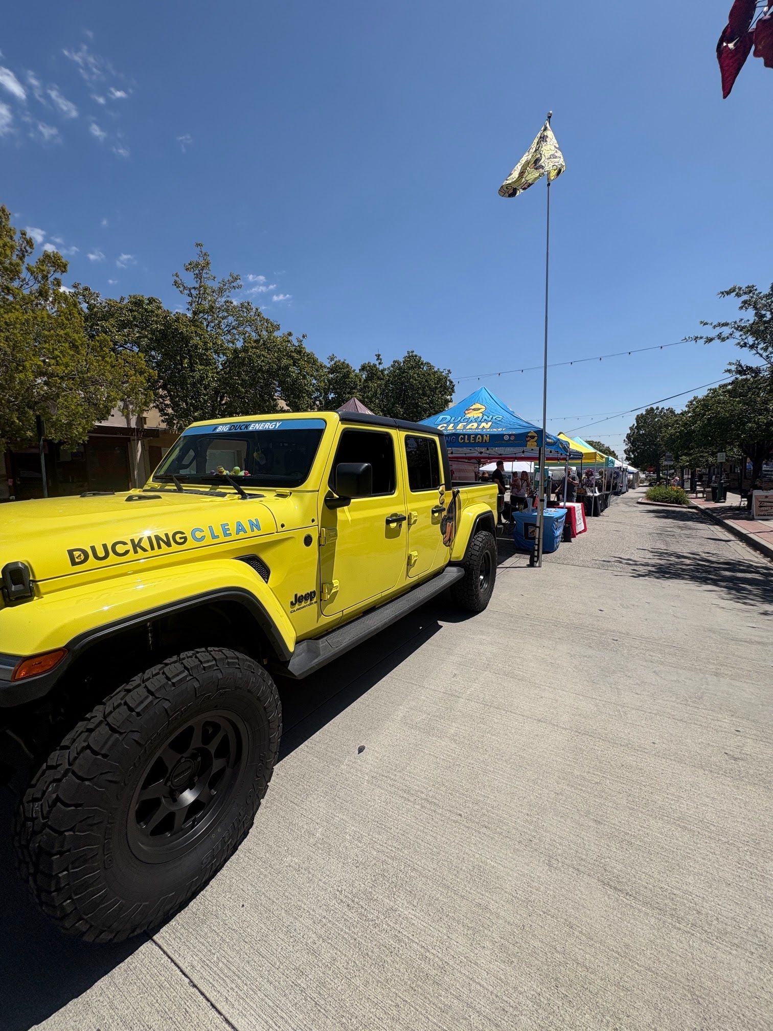 Yellow pickup truck parked beside a blue event tent on a sunny street, with flags overhead.