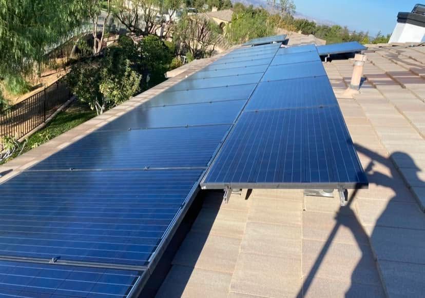 Blue solar panels installed on a sloped rooftop beside a beige tiled roof and trees