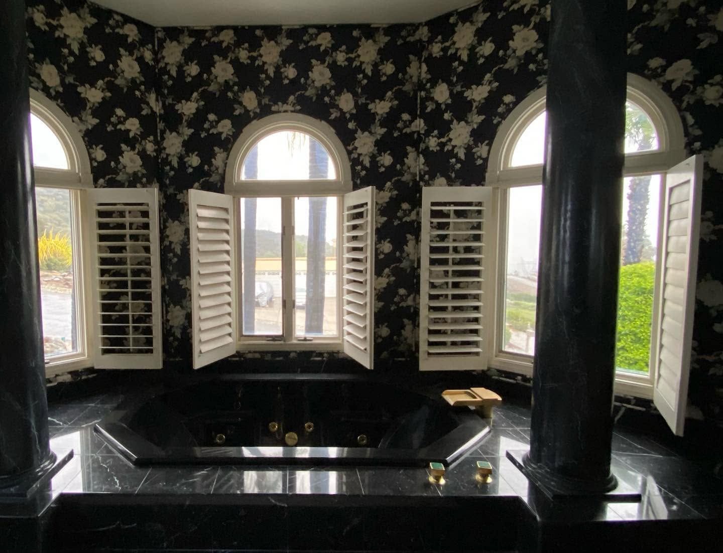 Black bathroom with floral wallpaper, arched windows, white shutters, and a glossy black vanity sink.