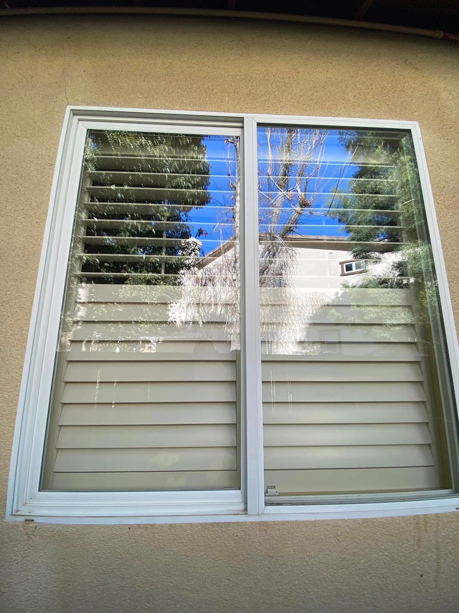 White-framed double window with blinds reflecting trees and blue sky on a beige exterior wall