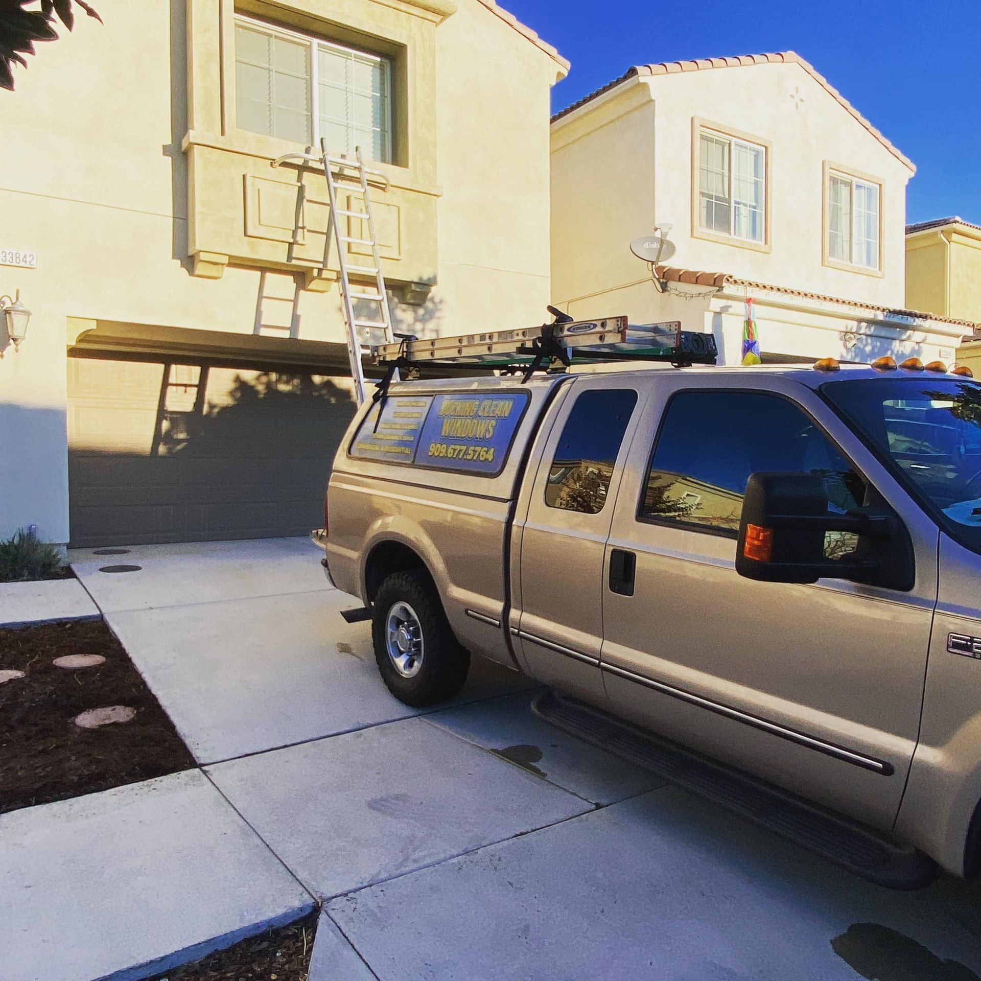 Silver work van in a driveway in front of a two-story house, with a ladder on the roof rack.