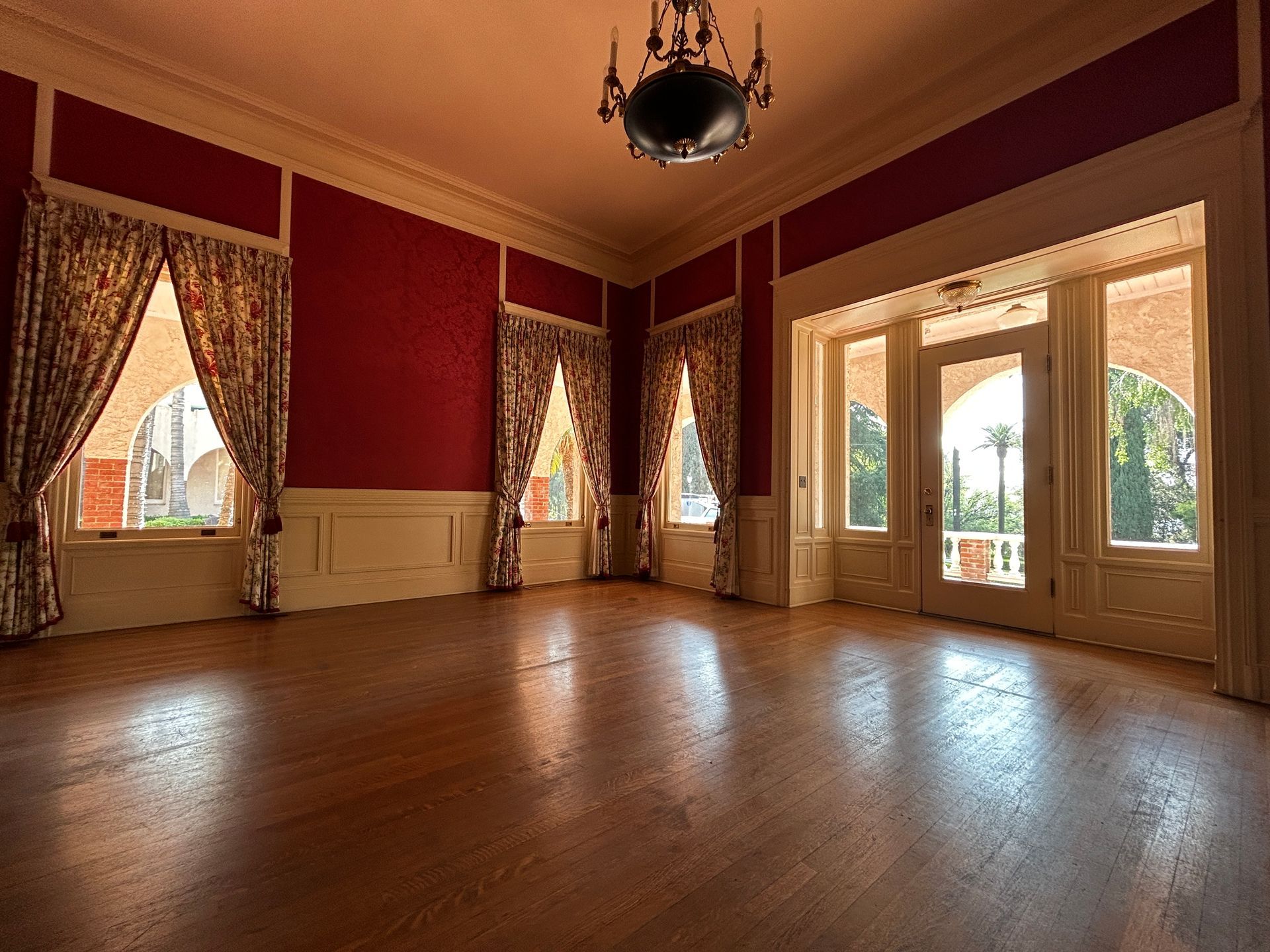 Empty elegant room with red walls, wood floor, tall windows, and chandelier.