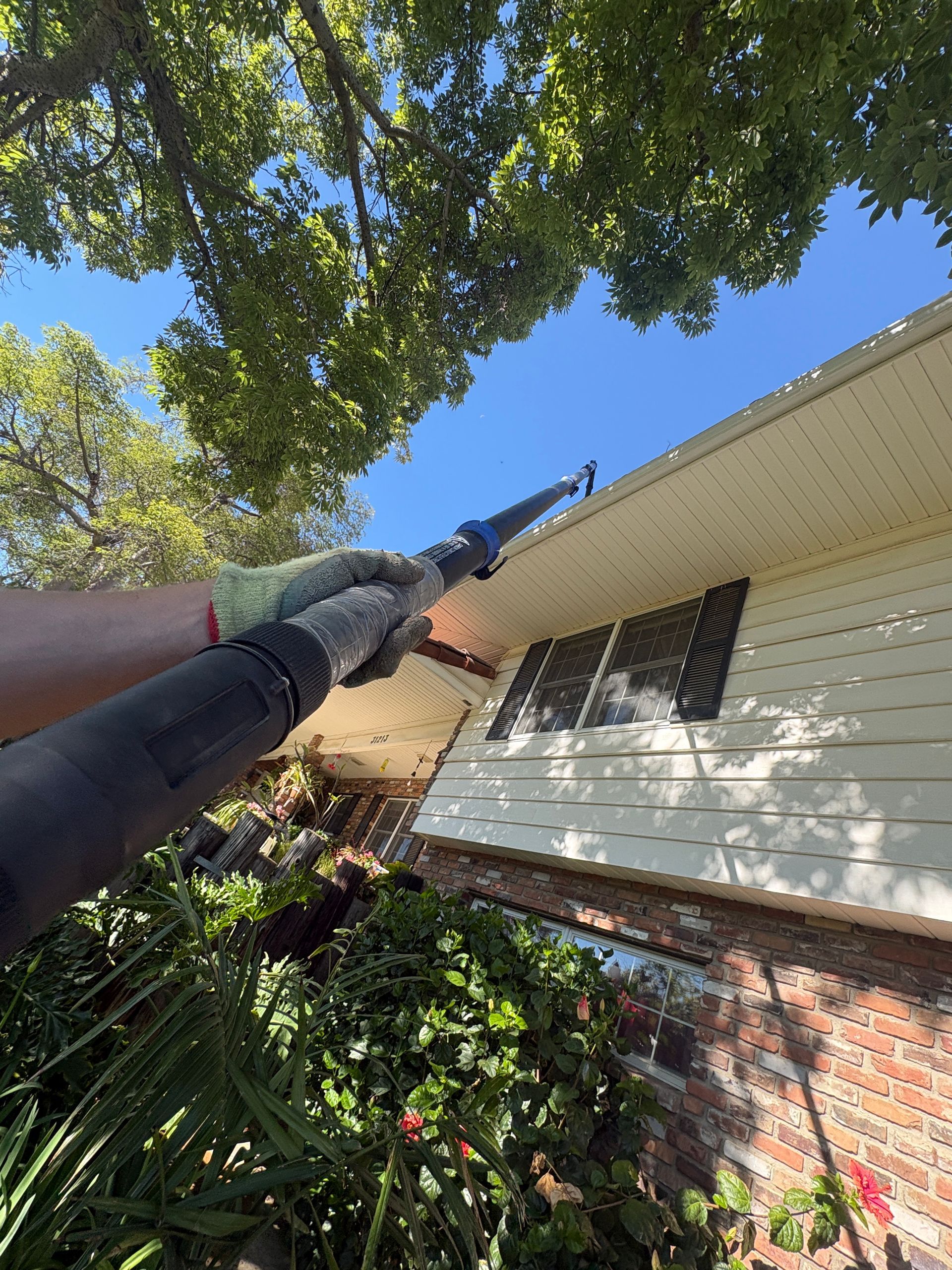 Gutter cleaning tool reaching a house roofline under tree branches on a sunny day