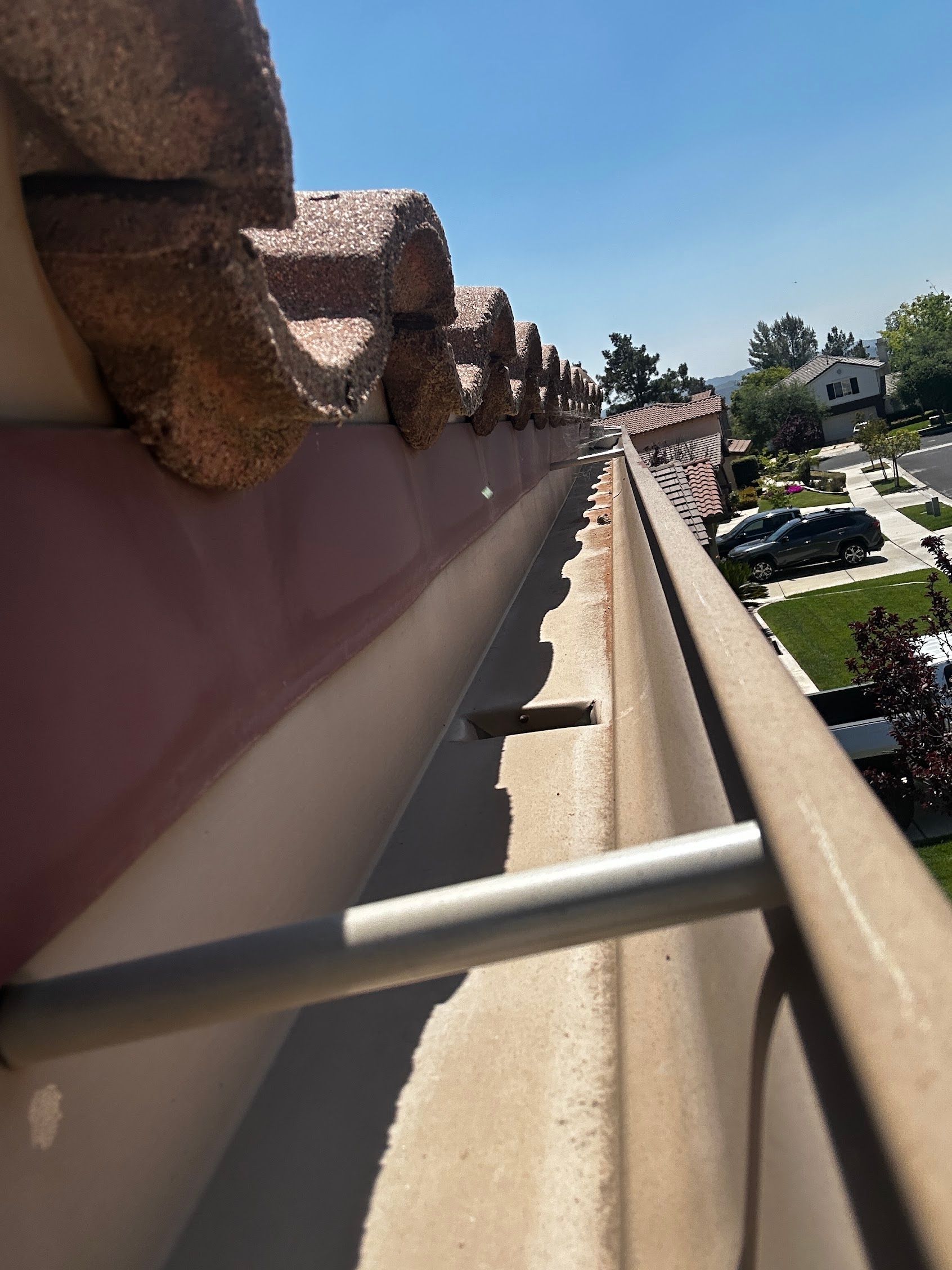 View down a sloped rooftop with terracotta tiles and a white metal railing, overlooking a parking lot and trees.