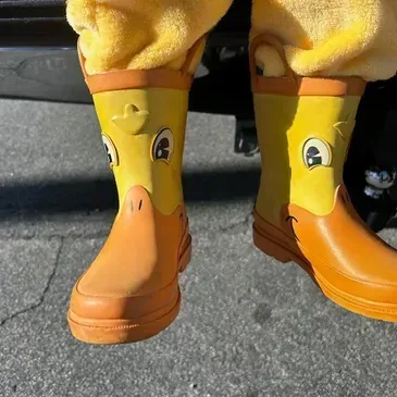 Yellow duck rain boots on a person standing on pavement, with cute duck faces and orange beaks.