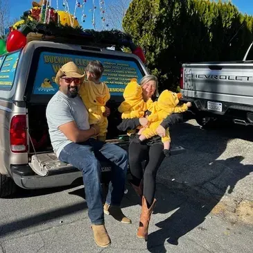 Two people sit on the tailgate of a pickup truck, holding yellow flowers in a sunny parking lot.