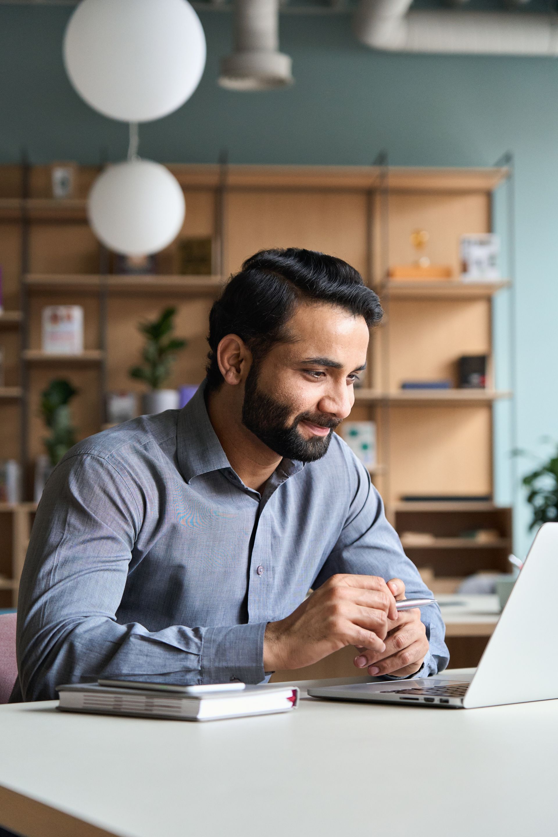 Man in a gray shirt smiling at a laptop at a desk in a bright home office