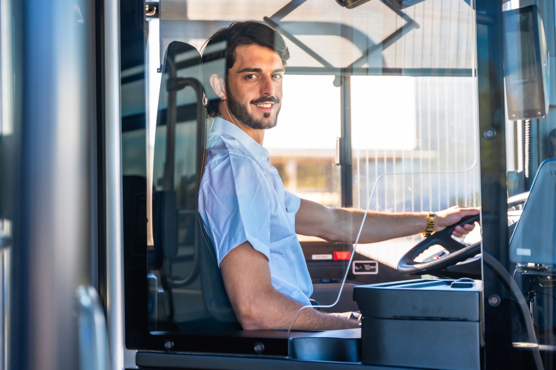 Man smiling in a bus driver’s seat, wearing a light blue shirt inside a vehicle cab