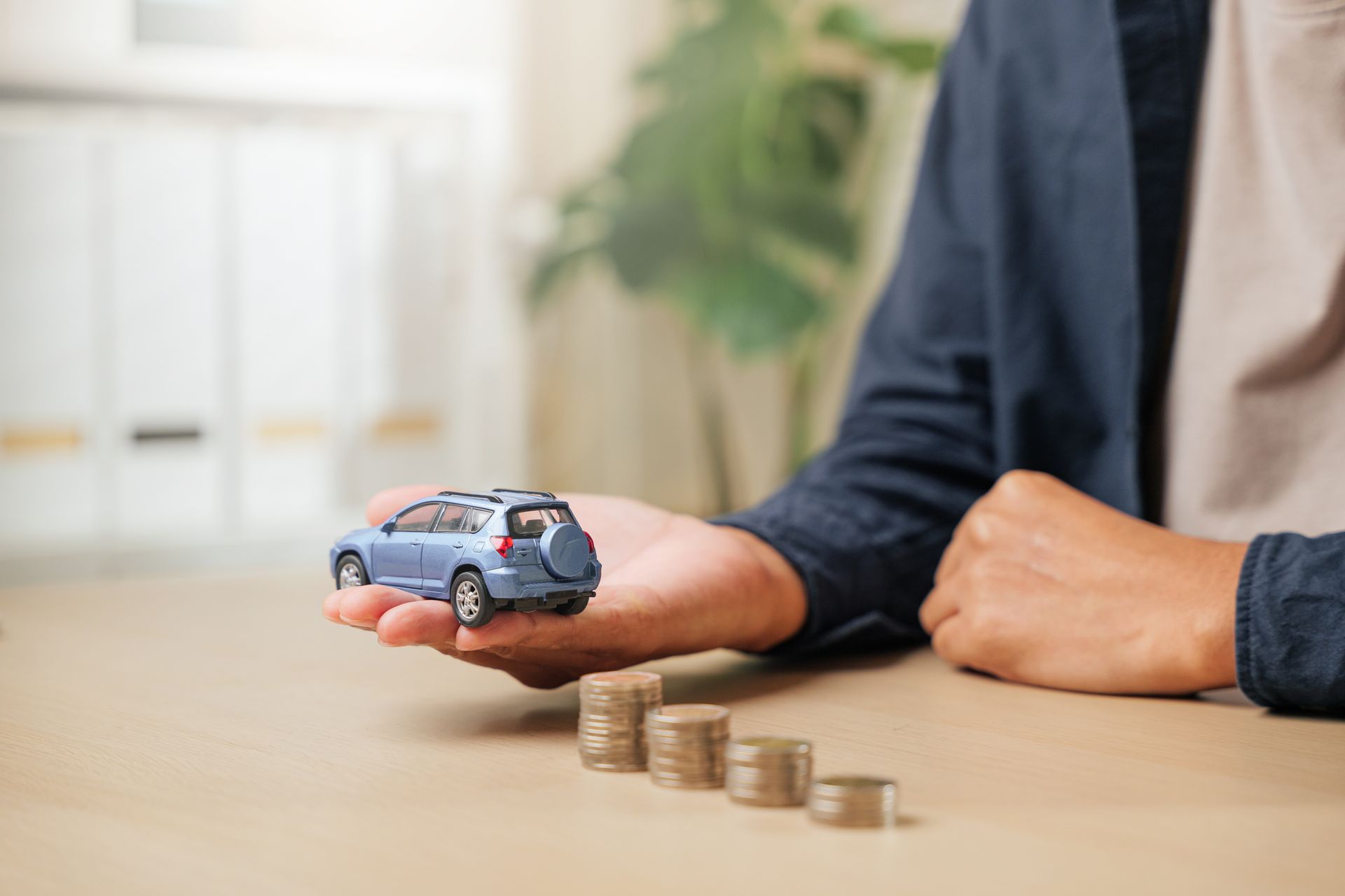 Hand holding a blue toy car beside stacked coins on a desk, suggesting car savings or insurance.