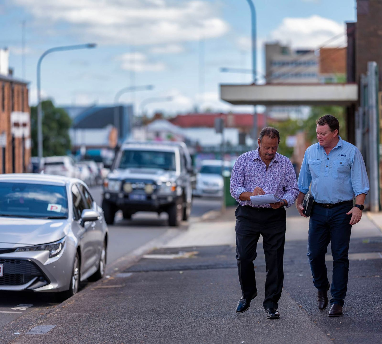Two men walking down a street, looking at paperwork. Cars and buildings in the background.