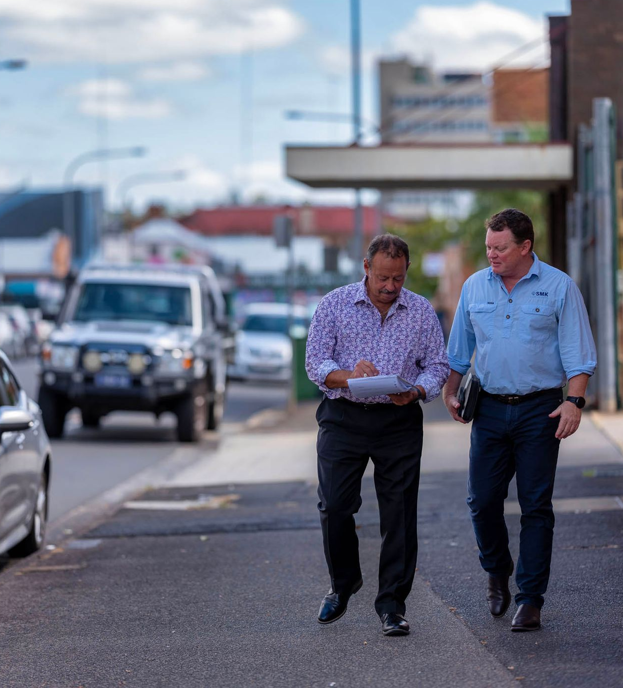 Two men walking down a street, one looking at papers. Cars and buildings are in the background.