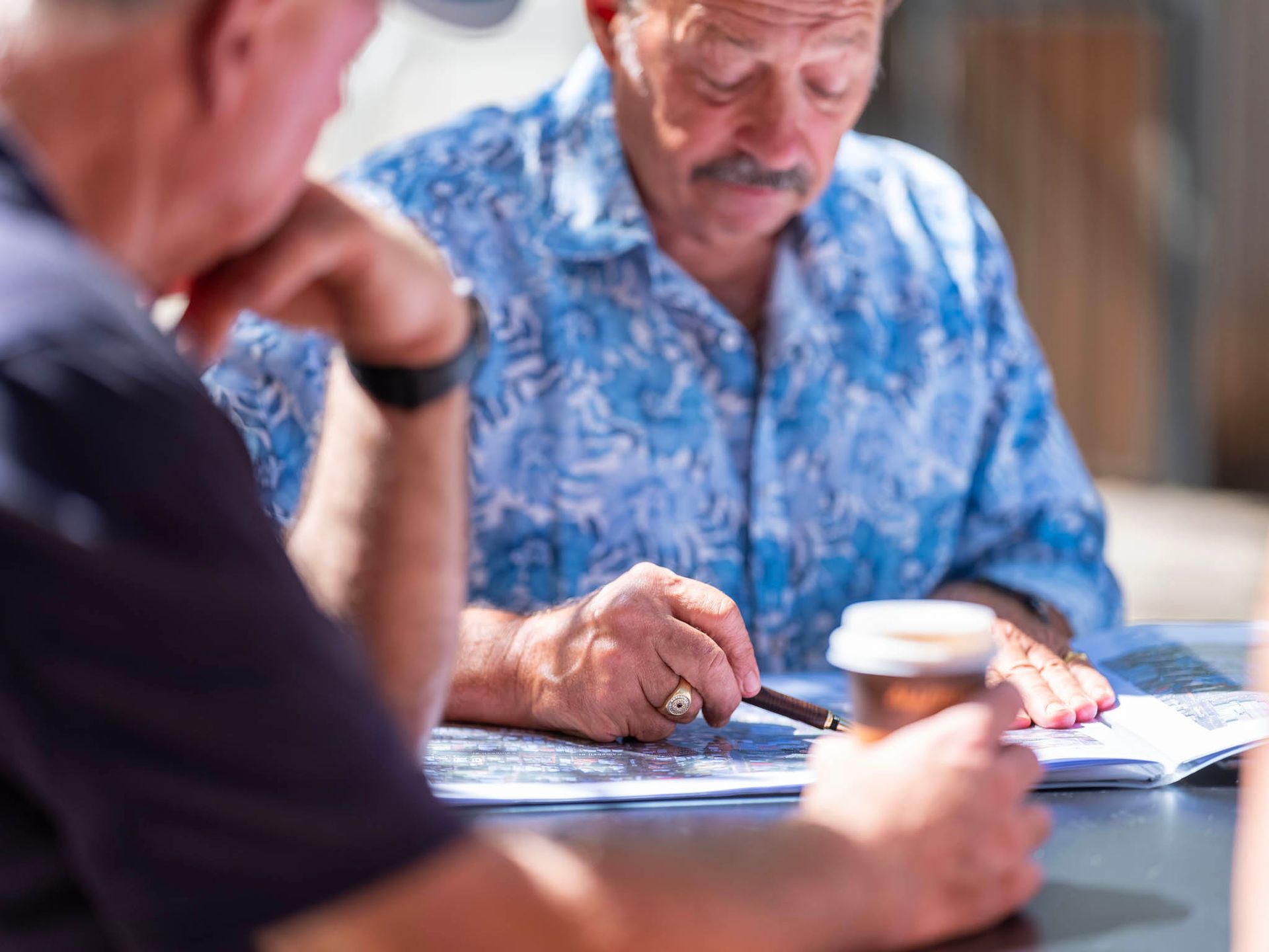 Two older men seated at a table, looking at papers. One points with a pen, the other holds a cup. Outdoor setting.