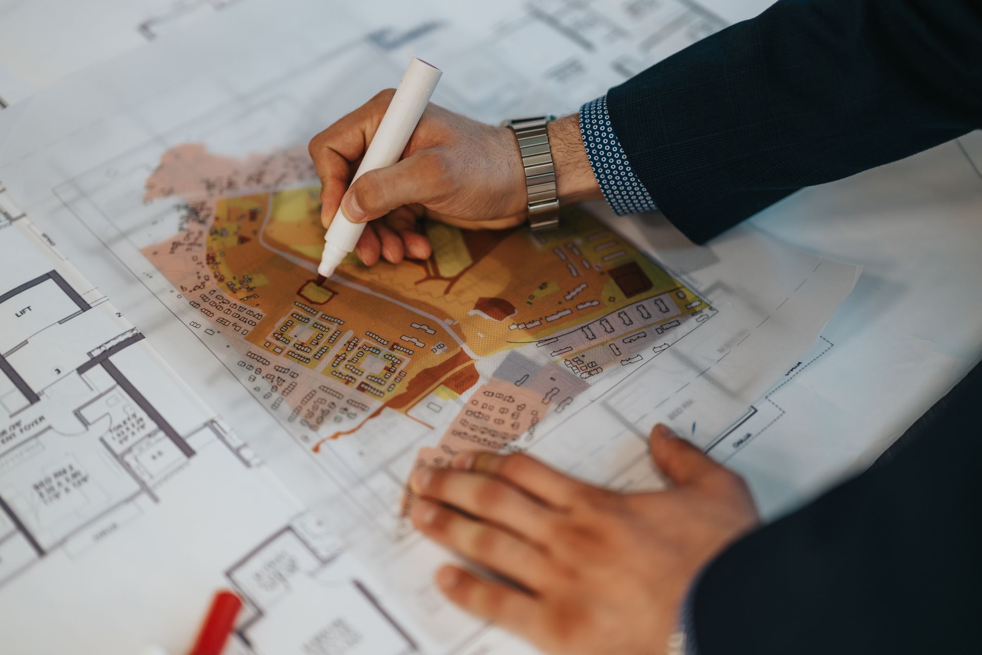 Person in suit jacket drawing on architectural blueprints with a white marker.
