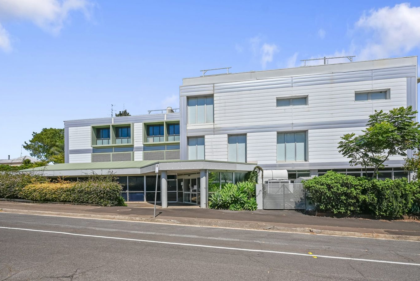 Gray and green multi-story building with large windows, bushes and a cloudy sky.