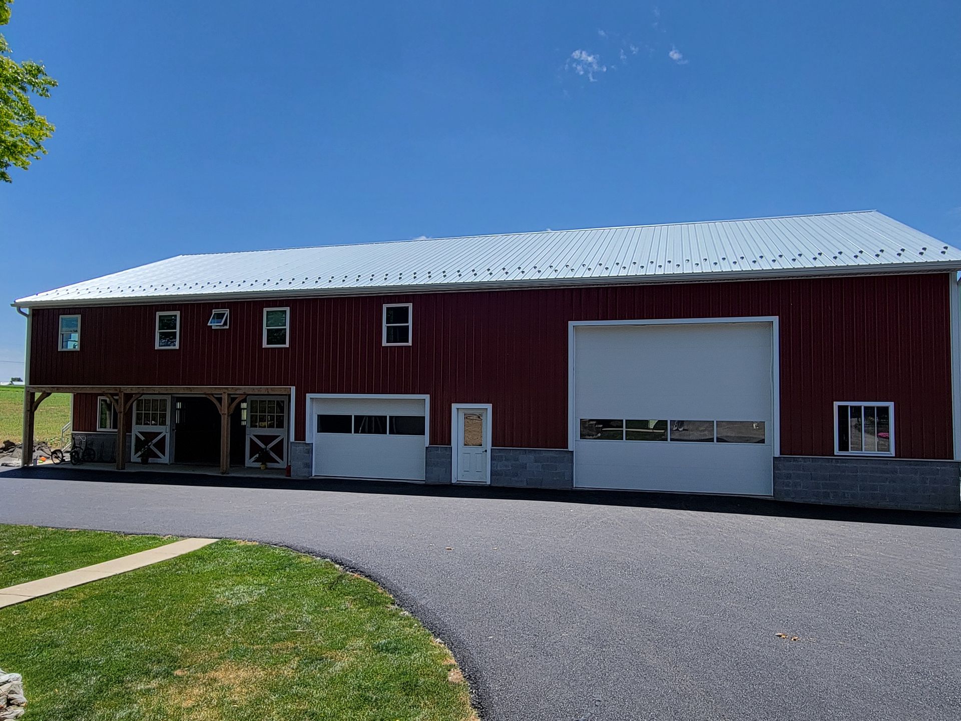 Red barn with white doors, metal roof, on a paved driveway, and grass lawn under a blue sky.