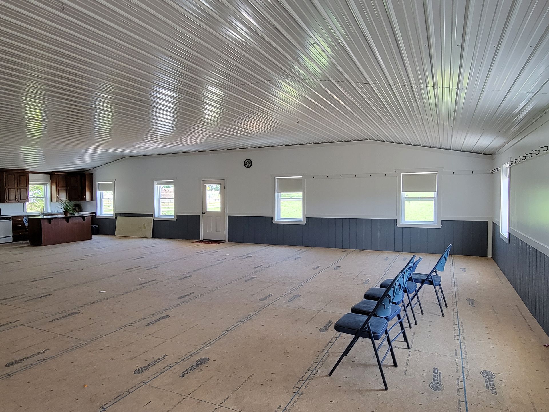 Empty, spacious room with corrugated metal ceiling. Chairs are lined up. Gray and white walls, windows.