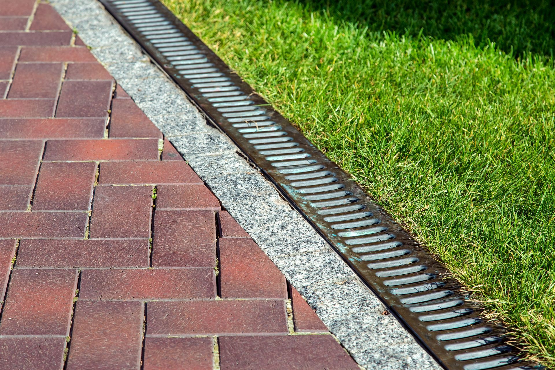 Brick walkway beside green grass with a metal drainage channel in between