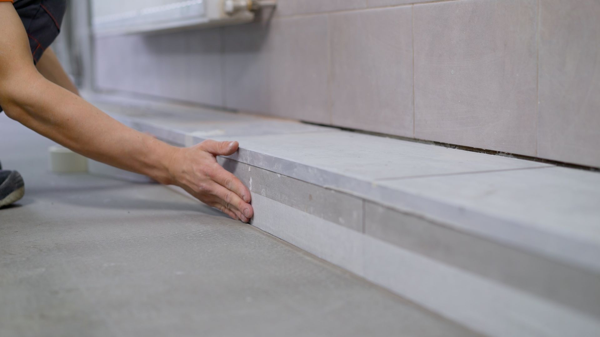 A person’s hand positions a light-colored tile piece against a low, tiered wall structure in an interior space.