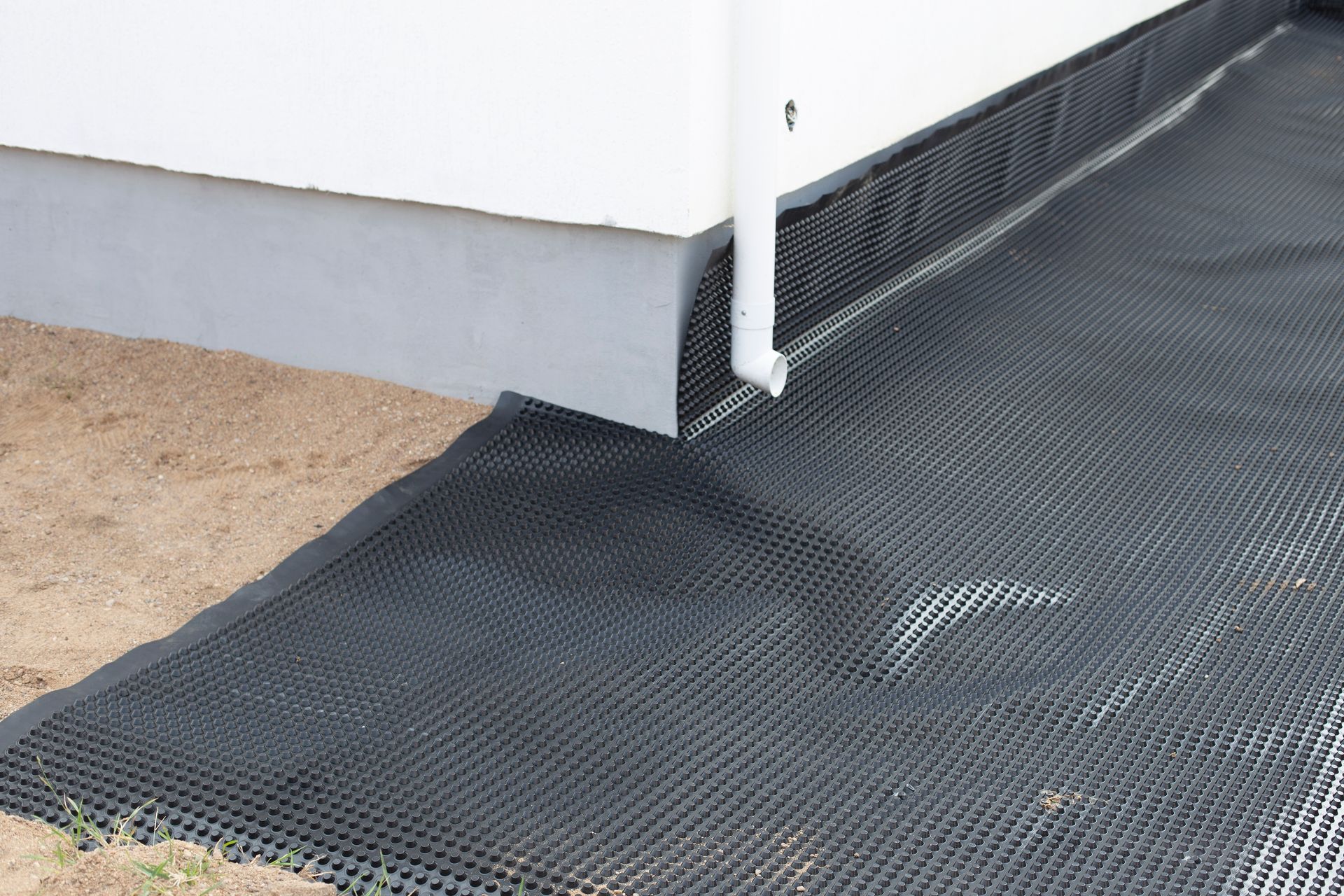 Black erosion control mat along a white building wall with a downspout and sandy ground nearby