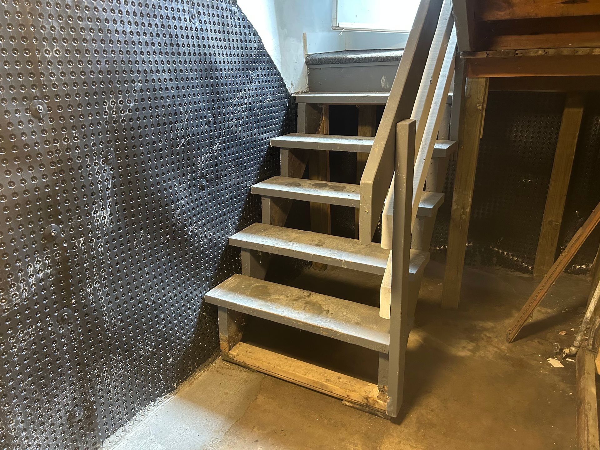 A short, grey staircase with wooden treads ascends against a textured, dark dimpled basement wall.