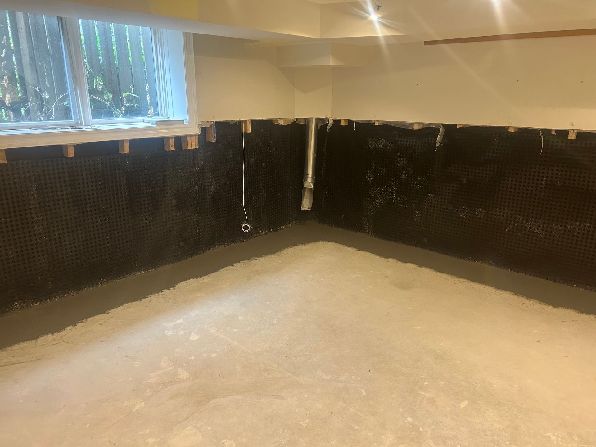 Basement corner with black mesh wall covering, gray concrete floor, window, and a white pipe.