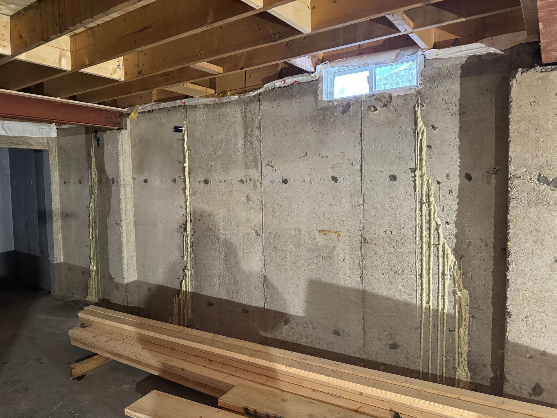 Basement interior with concrete wall, exposed ceiling beams, electrical wiring, small window. Wood planks on floor.