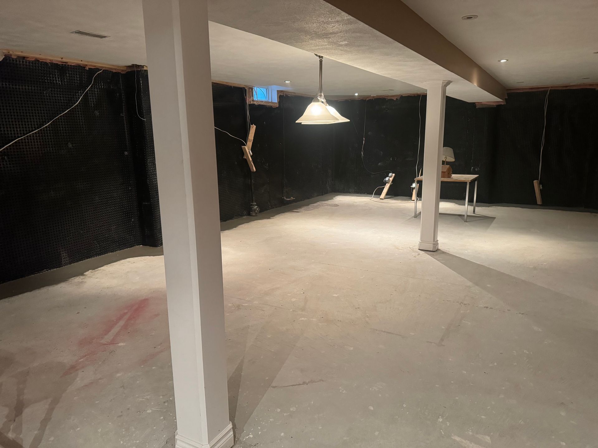 Basement interior with support beams, concrete floor, black insulation on walls, and a hanging light.