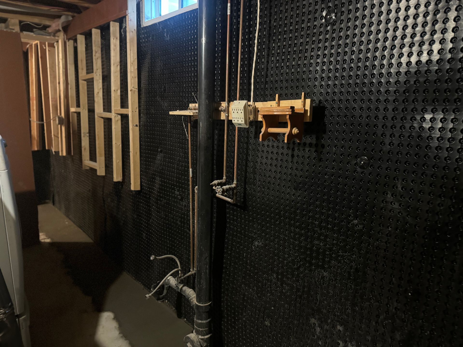 Basement wall with black dimpled membrane, pipes, and wooden framing.
