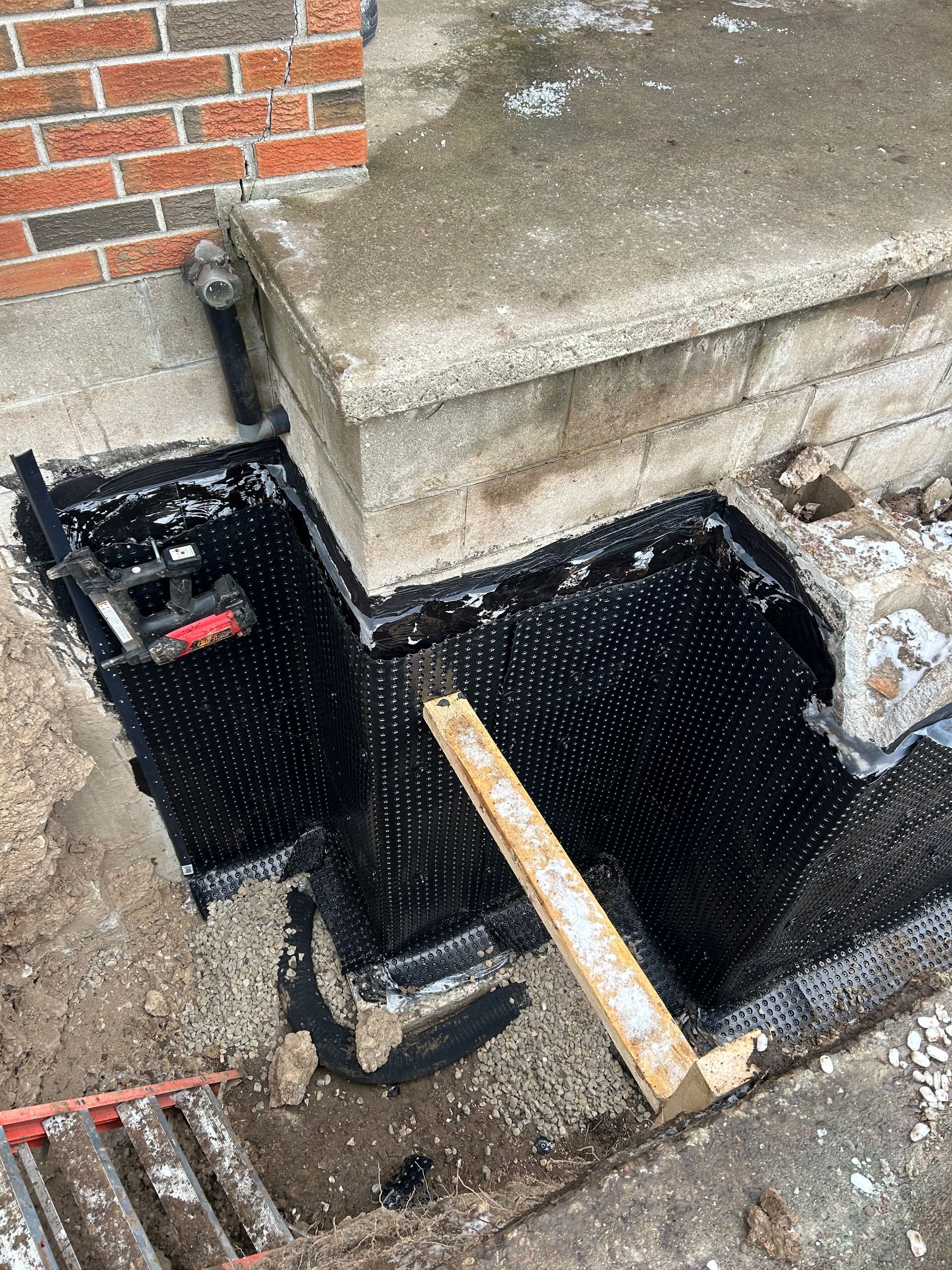 Construction site: a concrete step with a black mesh lining the excavated foundation. A wooden tool rests inside.